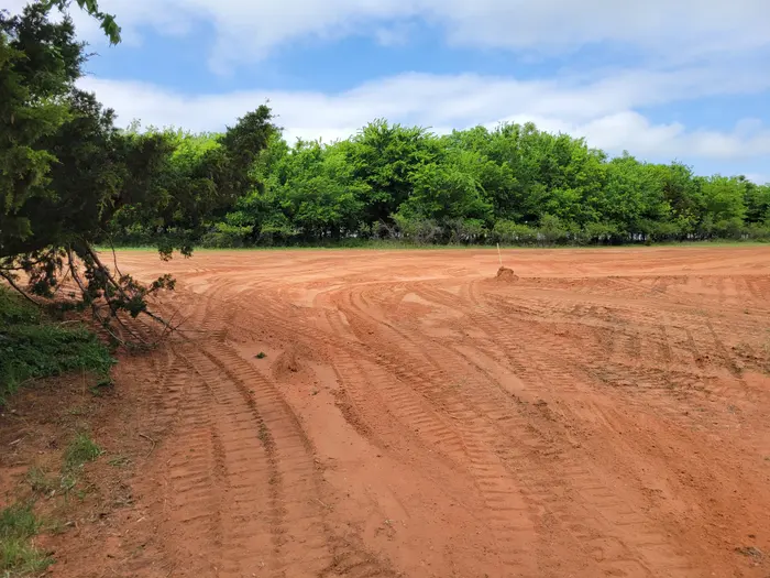 Cleared lot worked to bare red dirt, machine tracks across it, green tree line behind